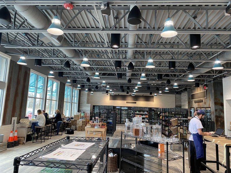 Sharp daylight wide shot of the finished Alltown Fresh market interior with dozens of pendant speakers and teal light fixtures across the truss ceiling, big storefront windows, and staff at work below.