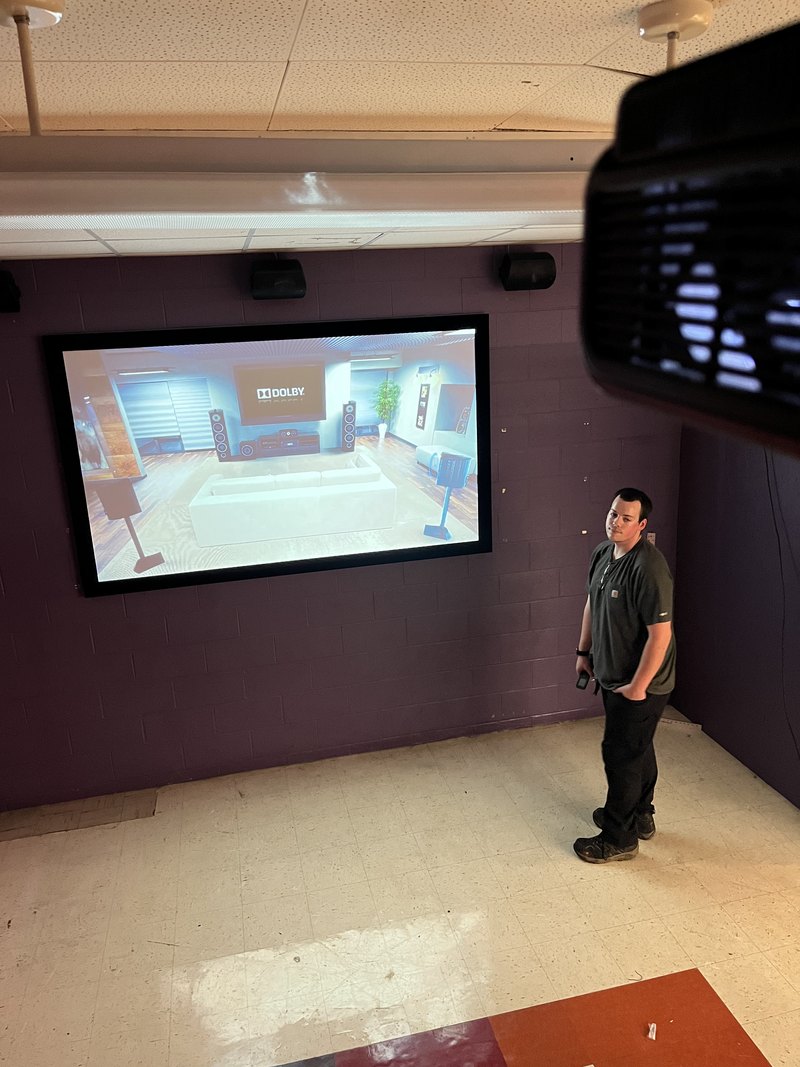 Finished media room with a large wall-mounted flat screen running a Dolby demo, flanked by wall-mounted surround speakers, as a tech looks on with remote in hand (EXIF-rotated).