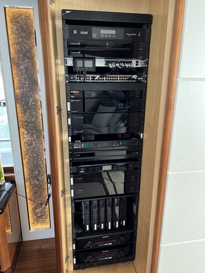 The finished rack installed flush in the salon's wood cabinetry above a backlit stone accent panel, with satellite receivers, Control4 controllers, network switch, matrix, tuners, zone amplifiers, and labeled DirecTV boxes neatly faced off.