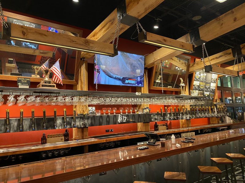Behind-the-bar panorama of the Lord Hobo tap wall and rows of racked glassware, with a wall-mounted TV playing between framed mirrors, chalkboard beer menus, and beam lights hung on chains.