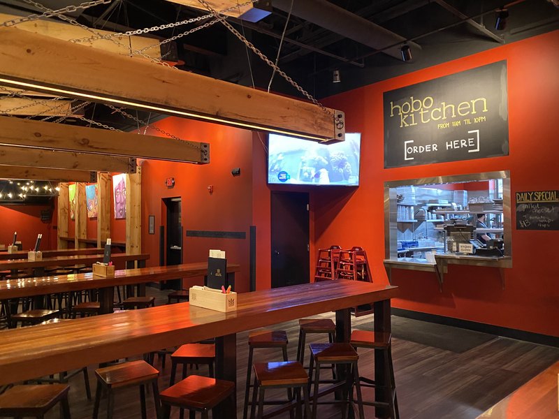 Wide taproom view with communal tables and stools under beam-integrated linear LED lighting, a corner-mounted flat-screen TV beside the Hobo Kitchen order window and chalkboard signage.