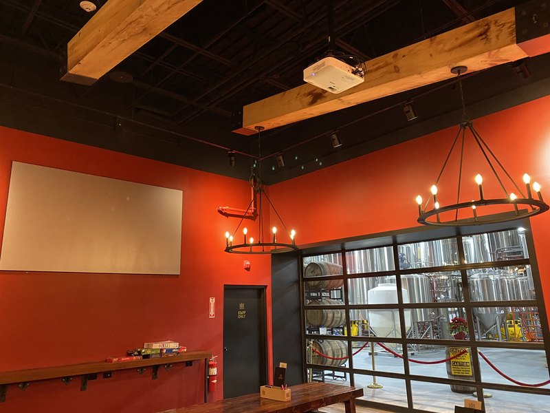 Event and game room with a wall-mounted projection screen and a beam-mounted Epson projector, ring chandeliers, and a divided-light window overlooking the stainless brewhouse and oak barrels.