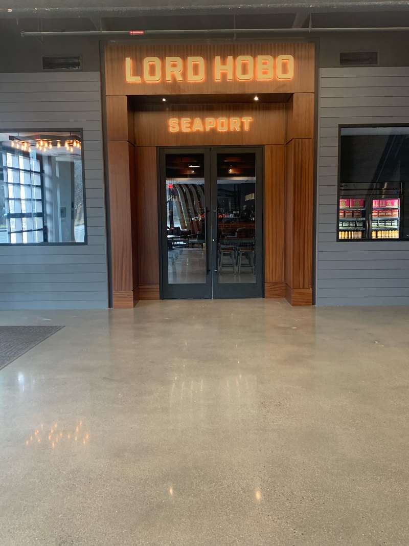 Lord Hobo Seaport entrance with halo-lit channel-letter signage over the wood-clad entry portal, accent downlights, and illuminated beer coolers visible through the window.