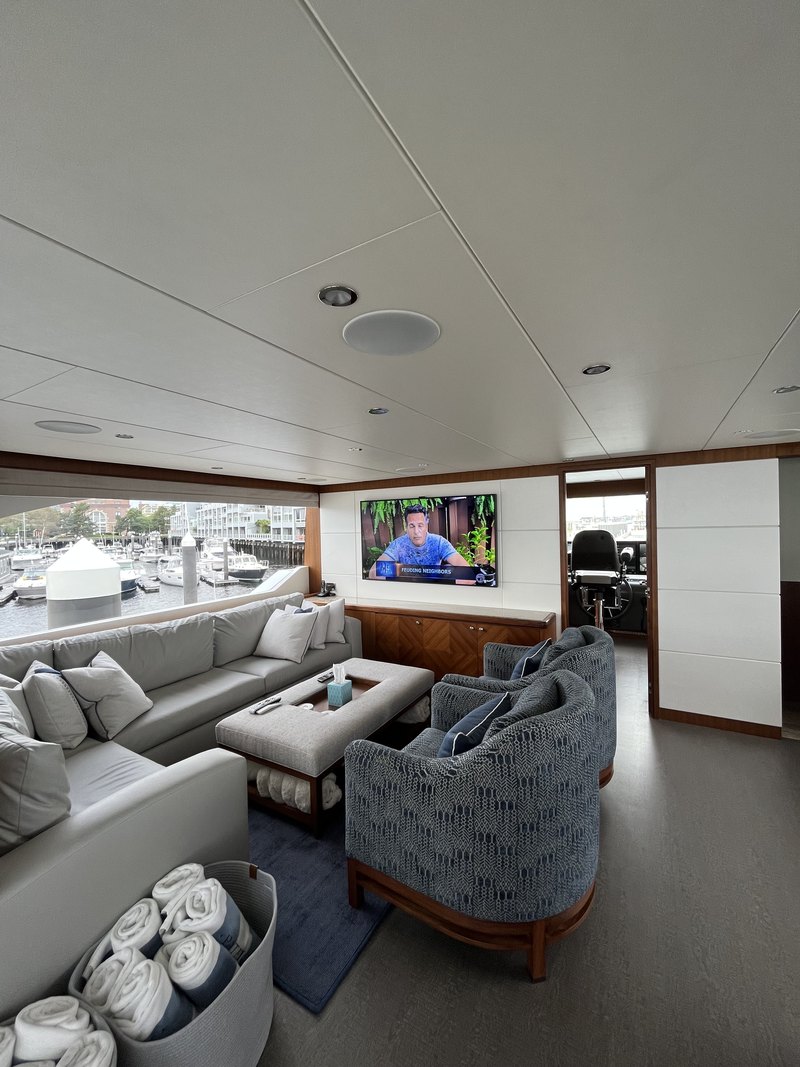 Wider salon view showing the wall-mounted TV in use, multiple in-ceiling speaker grilles and recessed downlights, sofa, and harbor view out the windows.