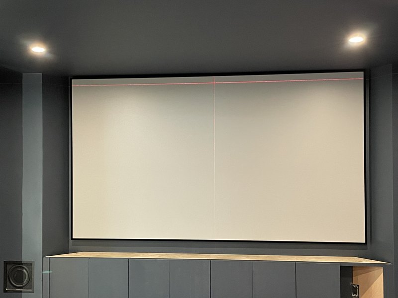 Finished screen wall: black fixed-frame projection screen mounted above gray equipment cabinetry with laser alignment line still glowing and recessed lighting on.