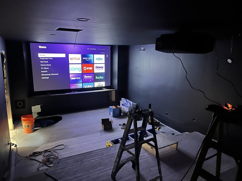 First light in the theater: fixed-frame screen glowing with the Roku home menu under a ceiling-mounted projector, ladders mid-install.