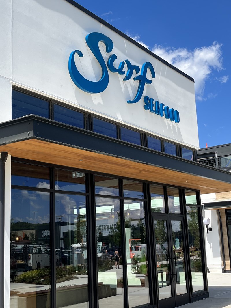 Exterior facade with the blue script 'Surf Seafood' channel-letter sign against a blue sky and glass storefront; content displays rotated 90 degrees and needs re-orienting, with a small exterior light and camera visible under the soffit.