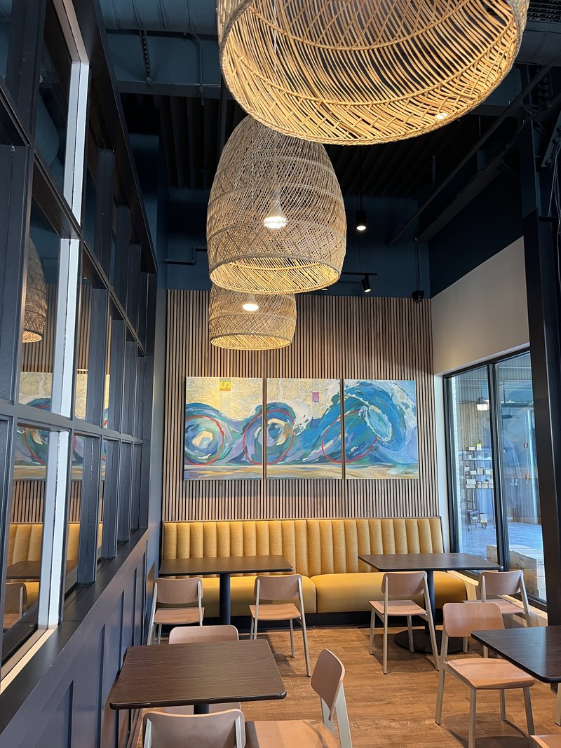 Dining room with rattan pendant light fixtures, mustard banquette seating, a wave-art triptych on a slat wall, and steel-framed glass partitions; content displays rotated 90 degrees and needs re-orienting before use.